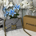 A gray PINKO shoulder bag with gold chain straps and a gold PINKO logo clasp, styled next to its brown cardboard box and decorative white and blue flowers against a patterned curtain backdrop.